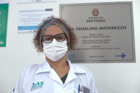 A foto traz Mônica, uma mulher jovem, de cabelos crespos e curtos, usando uma máscara, em frente a placa de identificação da unidade de saúde em que trabalha; na placa vê-se um brasão e o texto “Cidade de São Paulo” e “UPA Ermelino Matarazzo”  